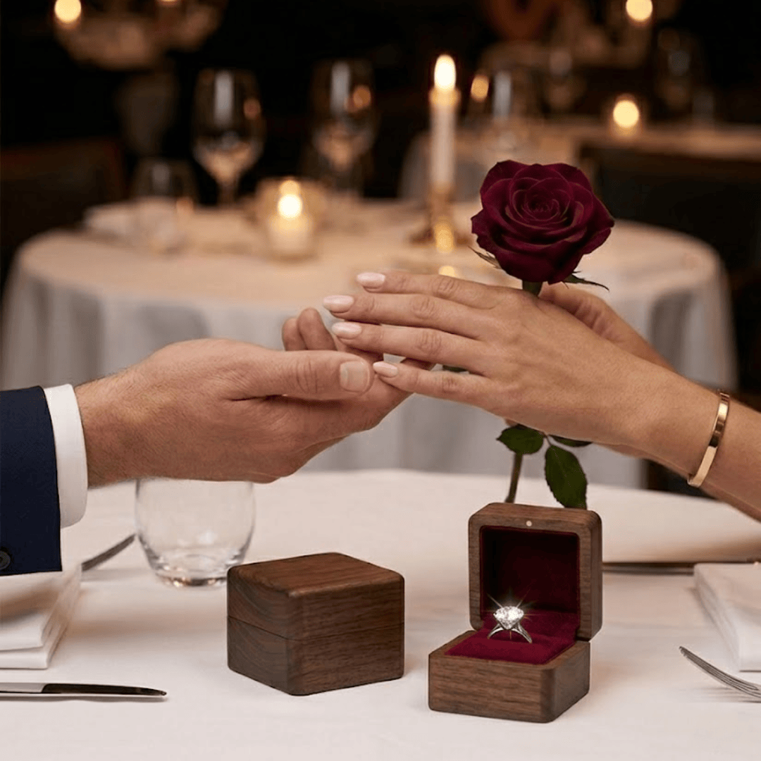 Gemini said A man and a woman hold hands across a restaurant table, with a diamond ring in an open wooden box in the foreground.