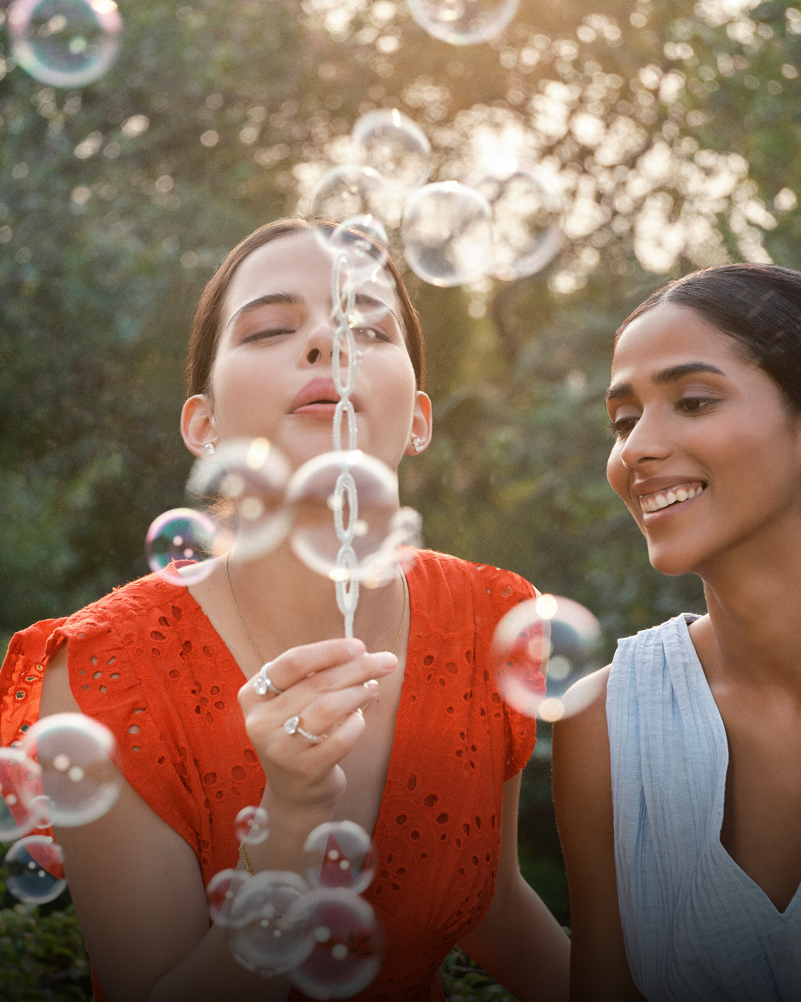 Two women smiling and blowing bubbles outdoors, wearing rings.